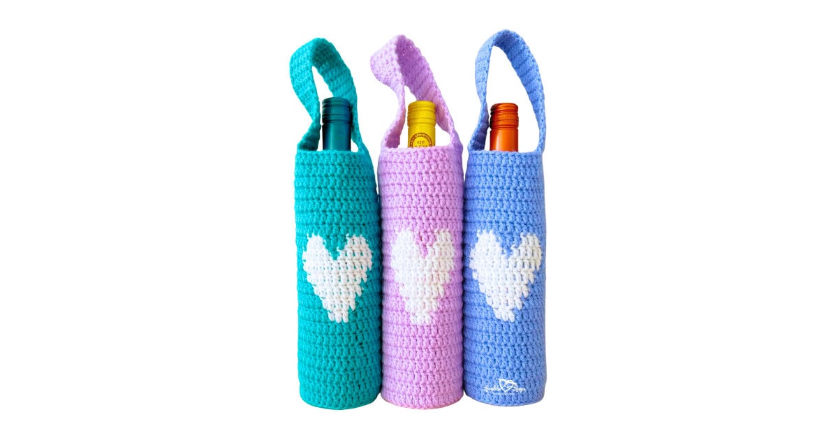 Three crochet wine bottle holders with straps in teal, pink, and blue, each with a white heart on the front&mdash;easy crochet wine bottle holder free pattern and reusable crochet wine bag gift.