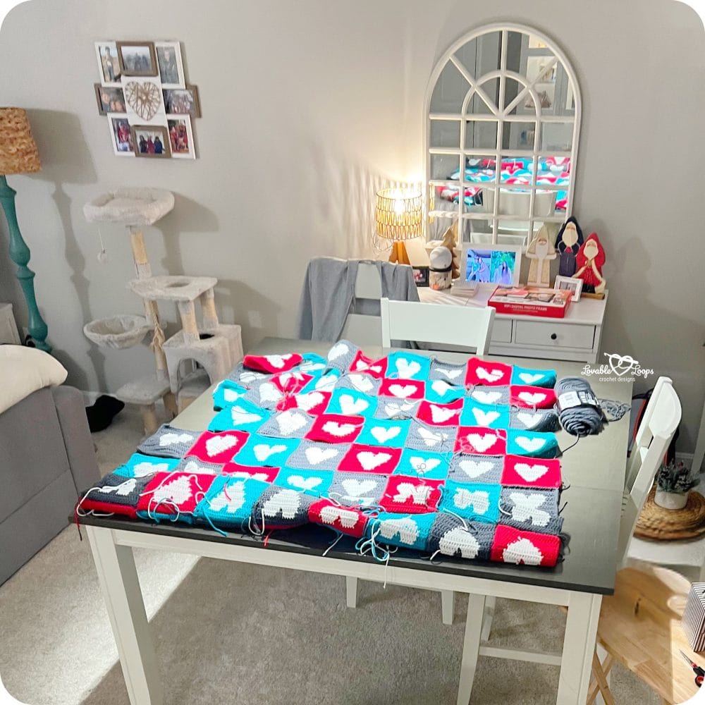 Crochet heart blanket squares laid out on a table before assembly, showing red, teal, and gray heart motifs made with double crochet stitches.