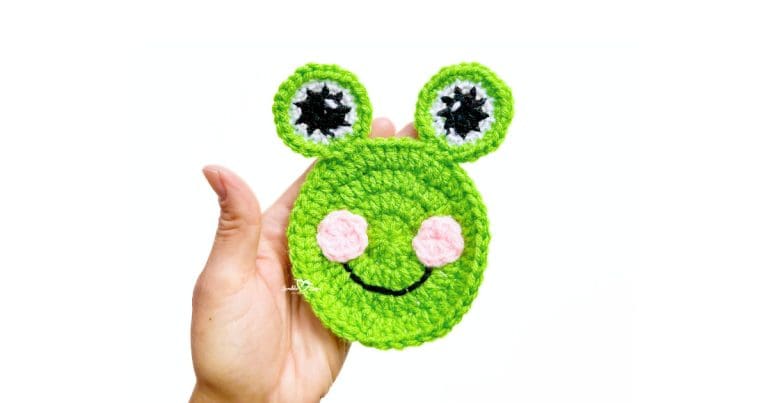Finished crochet frog applique held in hand, featuring a green frog face with pink cheeks, embroidered smile, and raised crochet eyes.