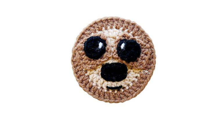 Finished crochet sloth applique made with brown and tan yarn, featuring stitched eyes, nose, and smile, shown on a white background.