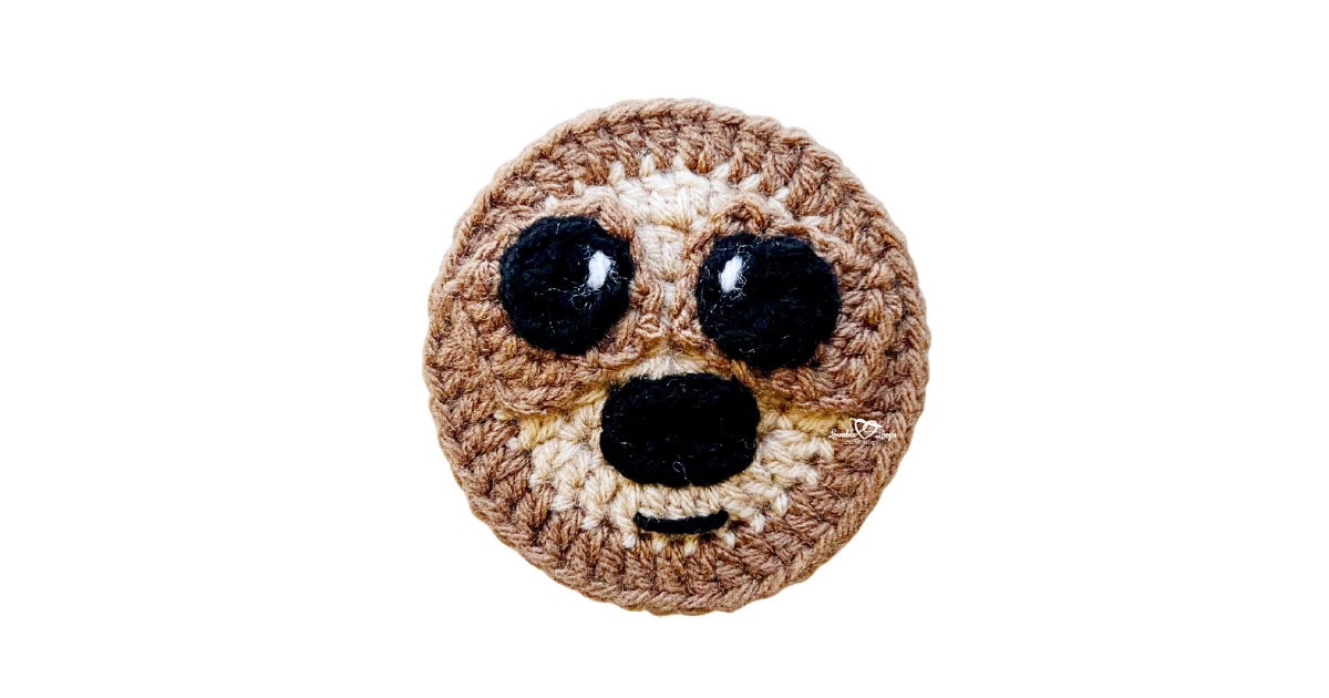 Finished crochet sloth applique made with brown and tan yarn, featuring stitched eyes, nose, and smile, shown on a white background.