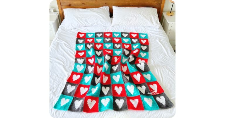 Handmade crochet heart blanket made from red, teal, and gray heart squares, arranged in a repeating pattern and displayed on a white bed as a cozy finished blanket.