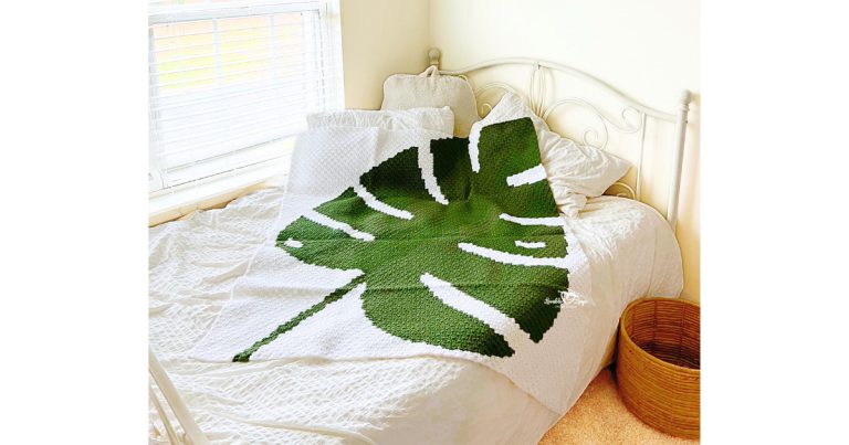 C2C crochet monstera leaf blanket draped across a white bed, showing a bold green monstera leaf design in a modern, neutral bedroom