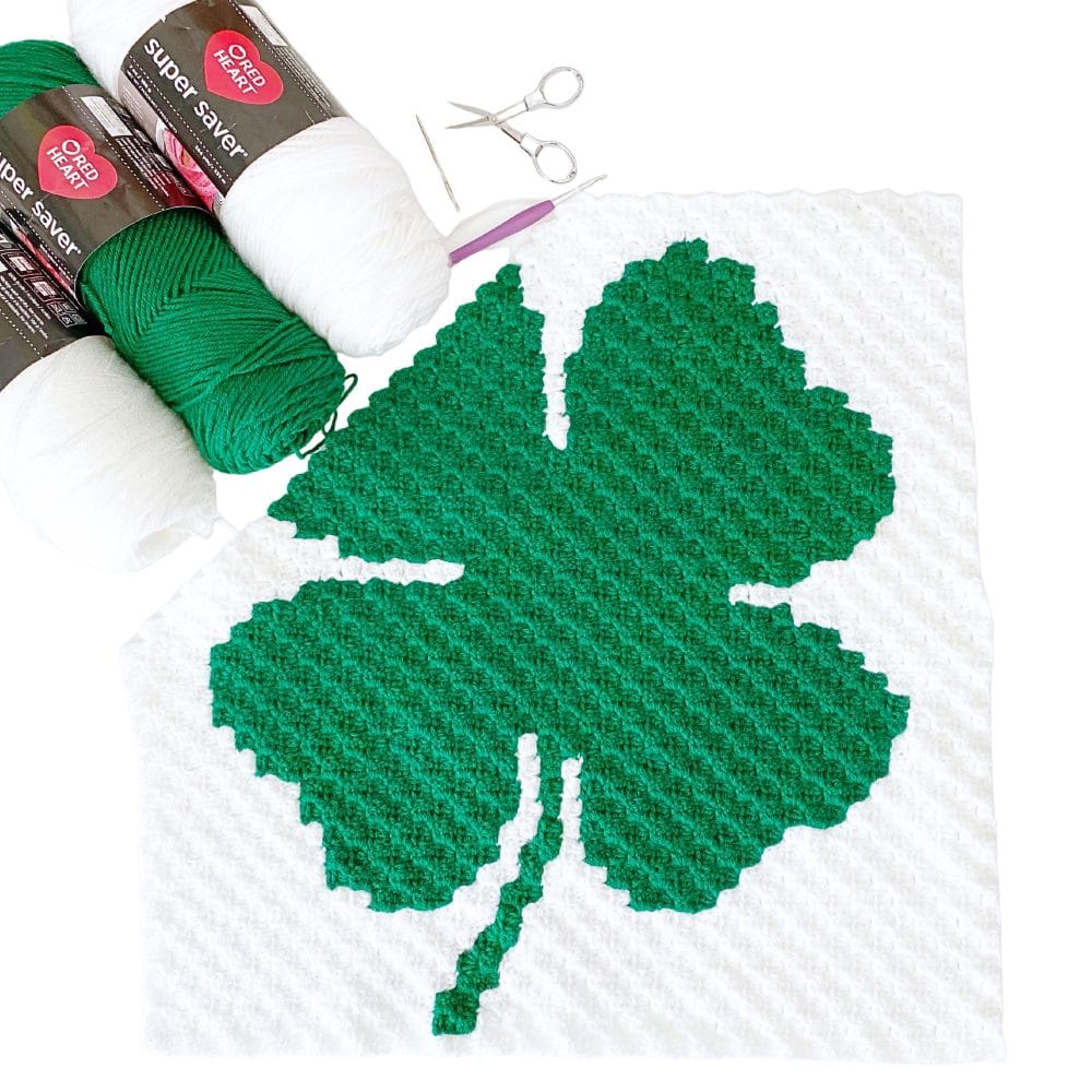C2C shamrock crochet pattern panel in green and white with worsted weight yarn, crochet hook, scissors, and yarn needle on a white background.