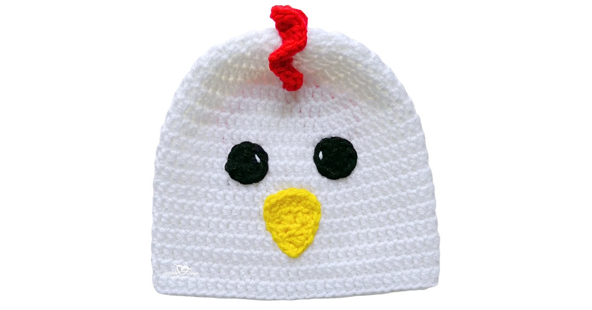 White crochet chicken hat with black eyes, a yellow beak, and a red comb on top.
