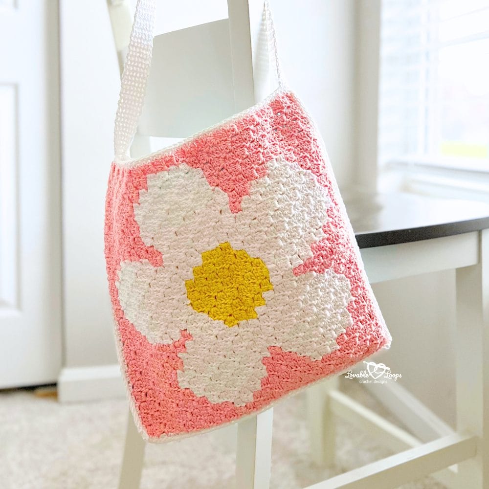 Crochet flower tote bag made with corner-to-corner crochet panels, featuring a white daisy with yellow center on a pink background and a white strap