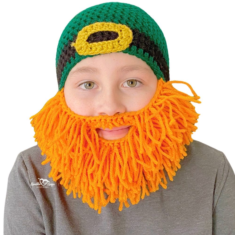Close-up of child wearing a crochet leprechaun beard hat pattern with green top-down beanie, black band, gold buckle, and bright orange yarn beard