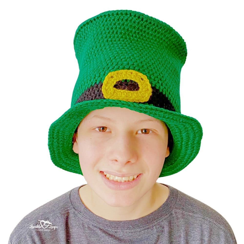 Close-up of a child wearing a tall green crochet leprechaun top hat with a black band and yellow buckle, perfect for St. Patrick&rsquo;s Day.