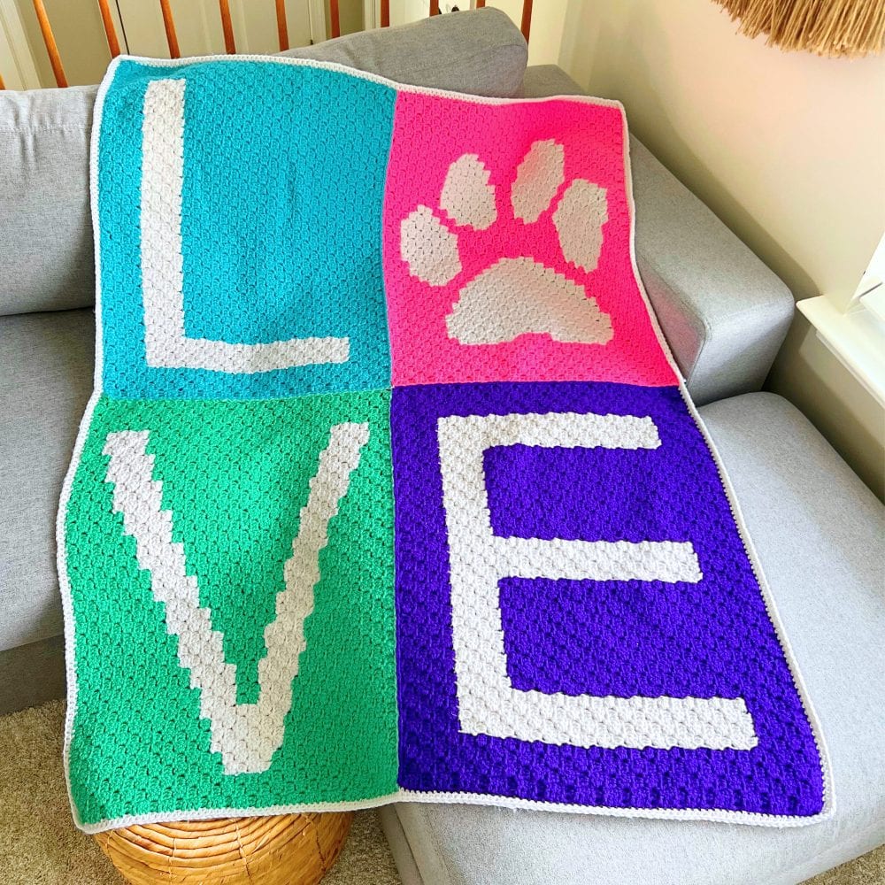 Corner-to-corner crochet love paw print blanket displayed on a couch, showing four colorful panels with a paw print and letter blocks in a C2C pet blanket pattern