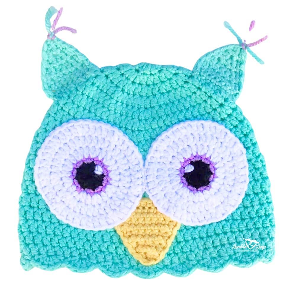 Detailed view of a crochet owl hat with large white eyes, purple accents, and a yellow beak attached to a teal hat