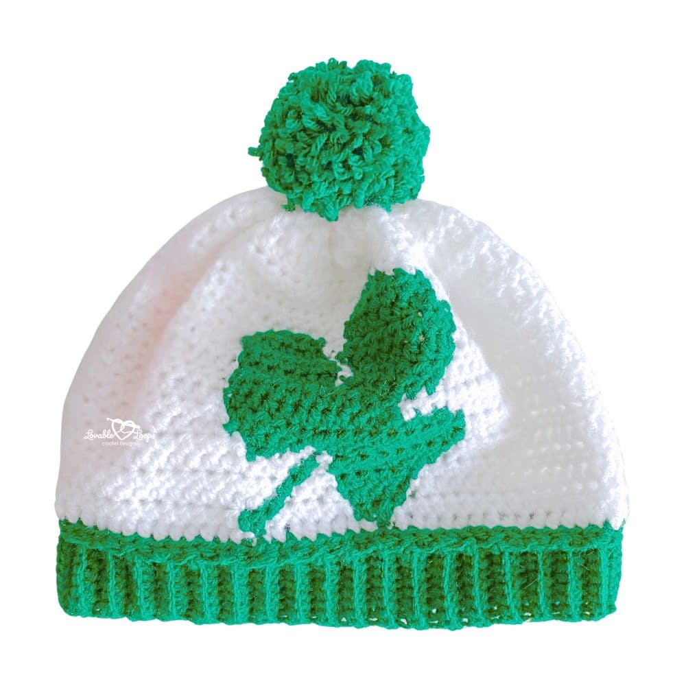 Close-up of finished crochet shamrock hat pattern featuring bold green shamrock, white hat body, ribbed brim, and green pom pom.