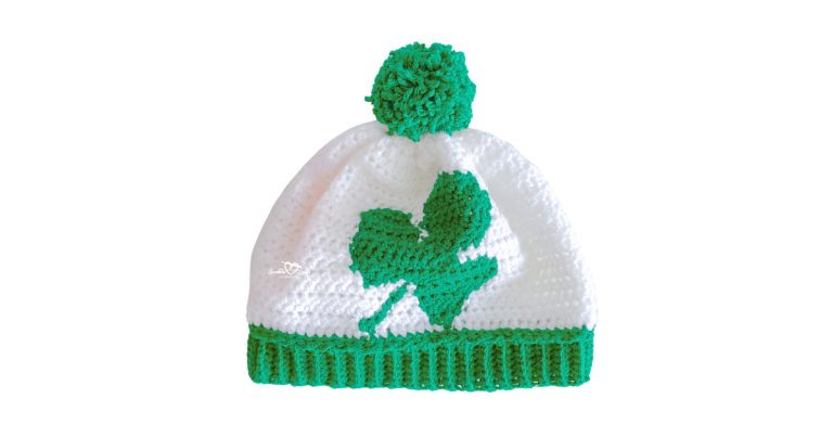 White crochet shamrock hat pattern with green shamrock design on front, green ribbed brim, and pom pom, worked flat in worsted weight yarn.