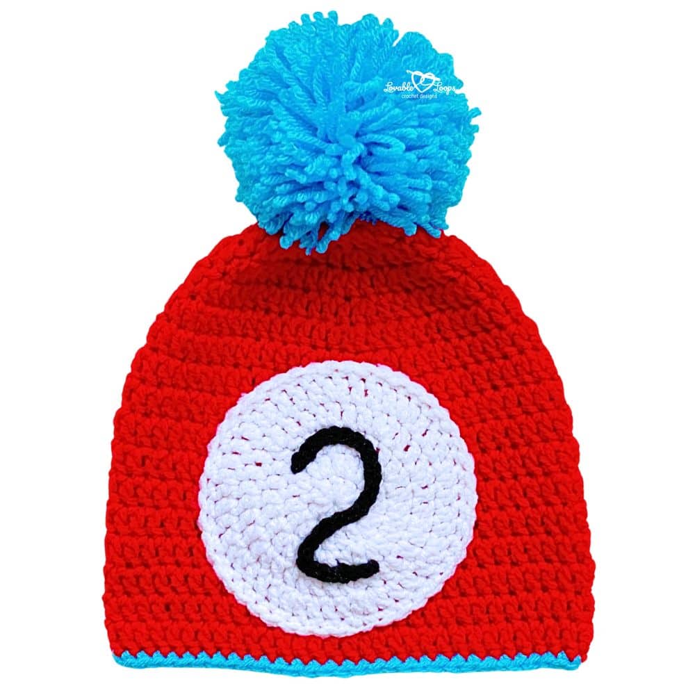 Finished red Read Across America crochet hat with white circle appliqué, black number 2, turquoise edging, and large blue pom pom.