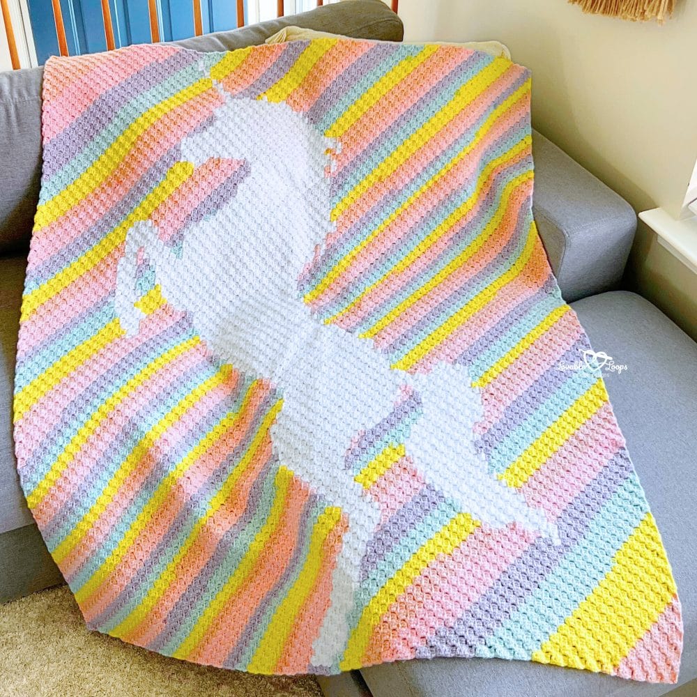 Corner to corner unicorn crochet blanket with pastel rainbow stripes and a white unicorn design, draped over a couch