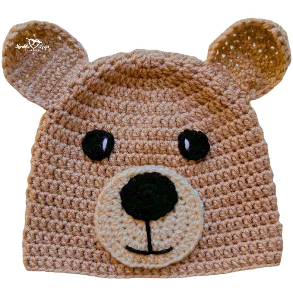 Close-up photo of a brown crochet bear hat with rounded ears, black safety eyes, and a cream snout with a black nose, shown on a light gray background.
