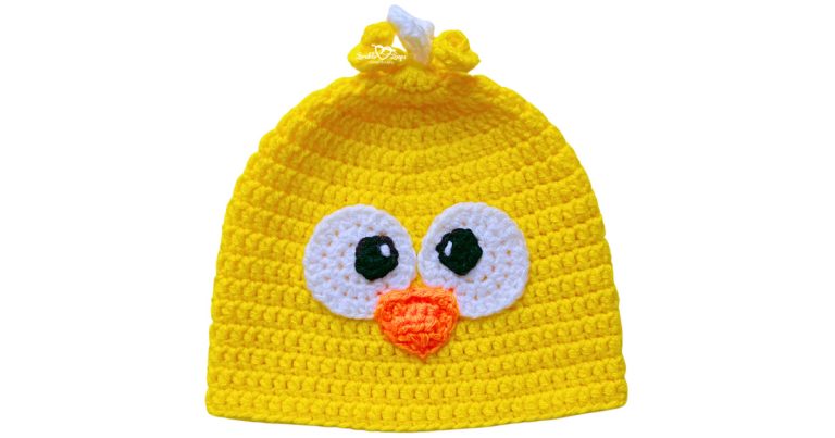 Bright yellow crochet chick hat with big white eyes, an orange beak, and a small white-and-yellow tuft on top.
