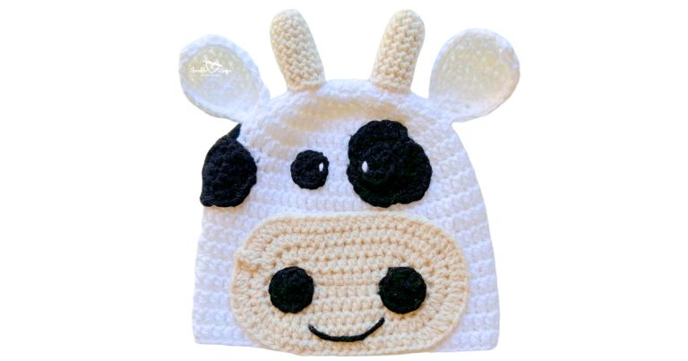 White crochet cow hat with black spots, tan horns, ears, and a large smiling muzzle on the front.