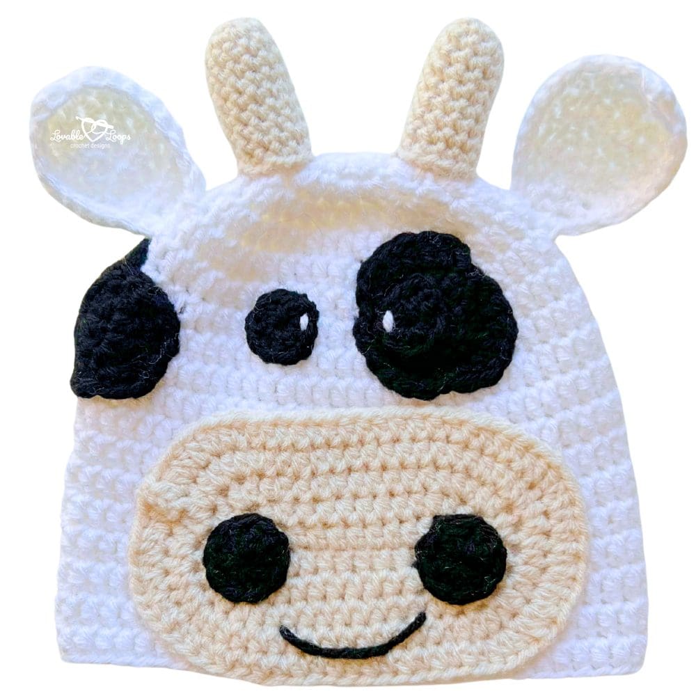 Handmade crochet cow beanie in white with black patches, soft horns, floppy ears, and a cheerful cow face.