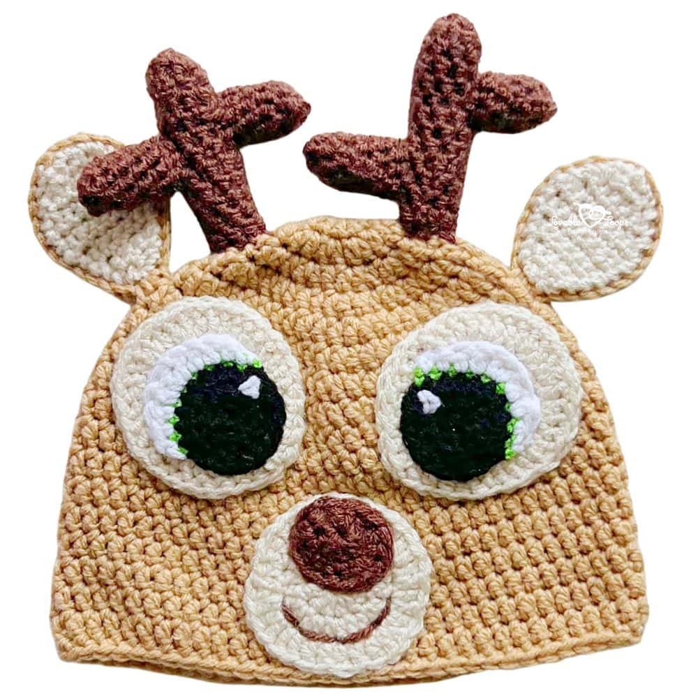 Close-up of a handmade crochet deer hat in tan and cream with brown antlers, ears, large eyes, and a round nose on a light background.