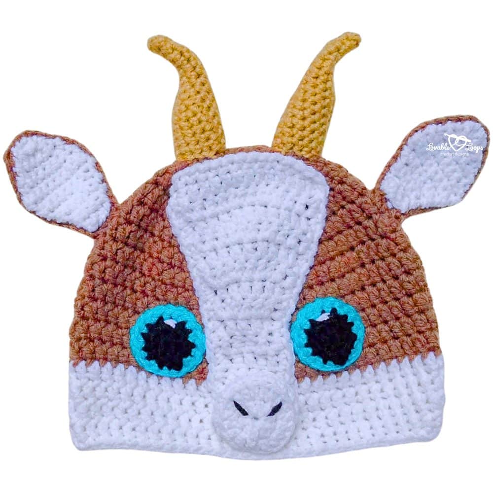 Close-up of a handmade crochet goat hat showing textured stitches, white snout, brown face, yellow horns, floppy ears, and bright blue eyes.