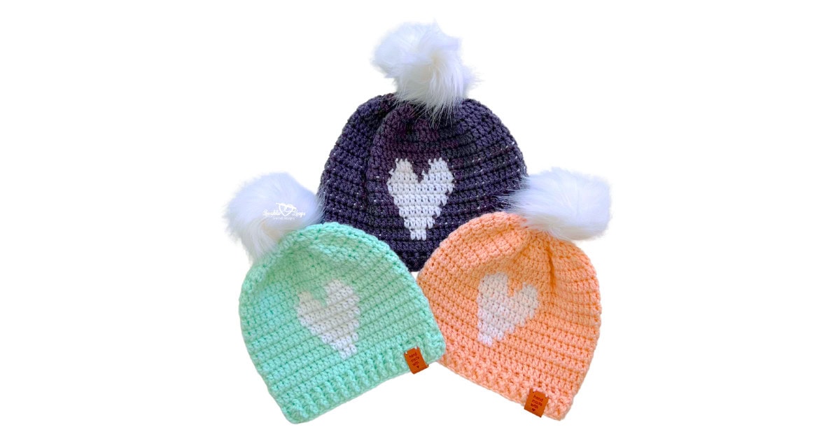Three handmade crochet heart beanies with white faux fur pom poms in mint, peach, and gray. Each beanie features a white heart design on the front. Free crochet heart beanie pattern includes three sizes for toddler, child, and adult.