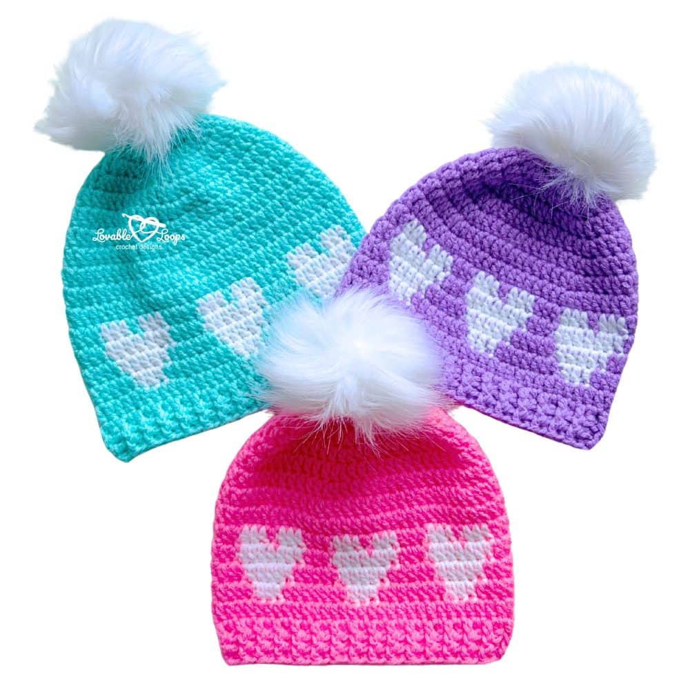 Pink, purple, and turquoise crochet heart beanies with white pom poms arranged flat. Easy crochet heart hat pattern for Valentine&rsquo;s Day in three sizes with heart rows and ribbed brim.