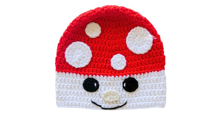Red and white crochet mushroom hat with white spots, black eyes, and a smiling face on the brim.