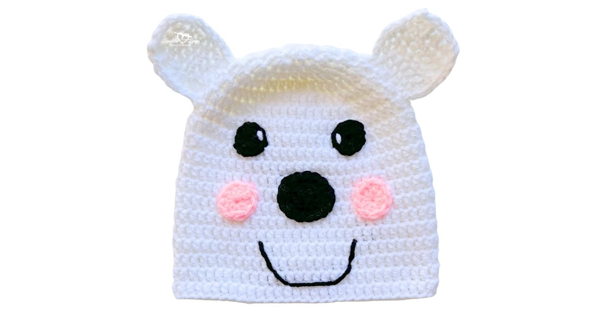 White crochet polar bear hat with round ears, black nose, pink cheeks, and a smiling face.