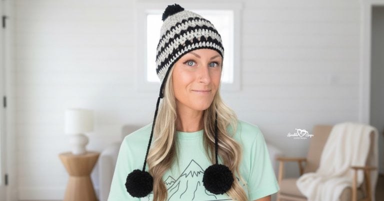 Woman wearing a black and white crochet pom pom beanie and mint green shirt, standing in a bright neutral living room with soft natural light.