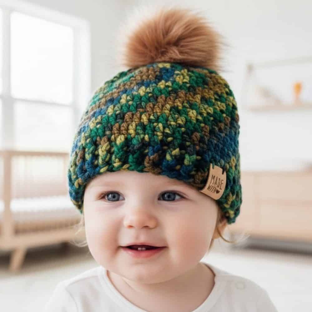 Baby wearing a multicolored crochet beanie in green, blue, and brown with a large faux fur pom-pom in a bright nursery.