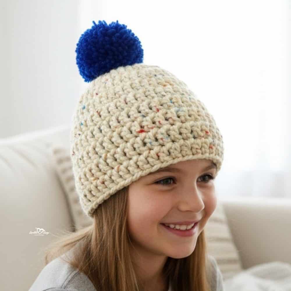 Smiling girl wearing a cream speckled crochet hat with a blue pom-pom in a bright neutral room.