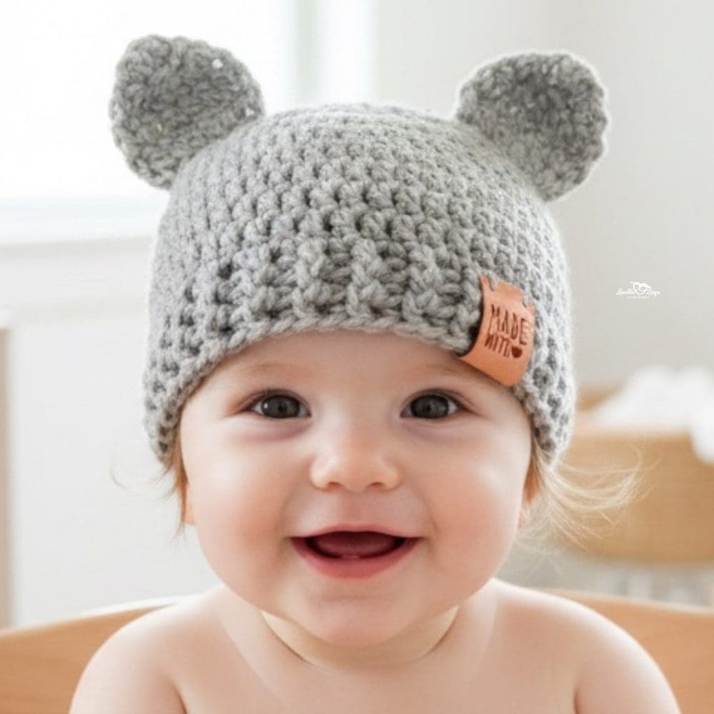 Smiling baby wearing a light gray crochet hat with small rounded ears in a bright neutral nursery.