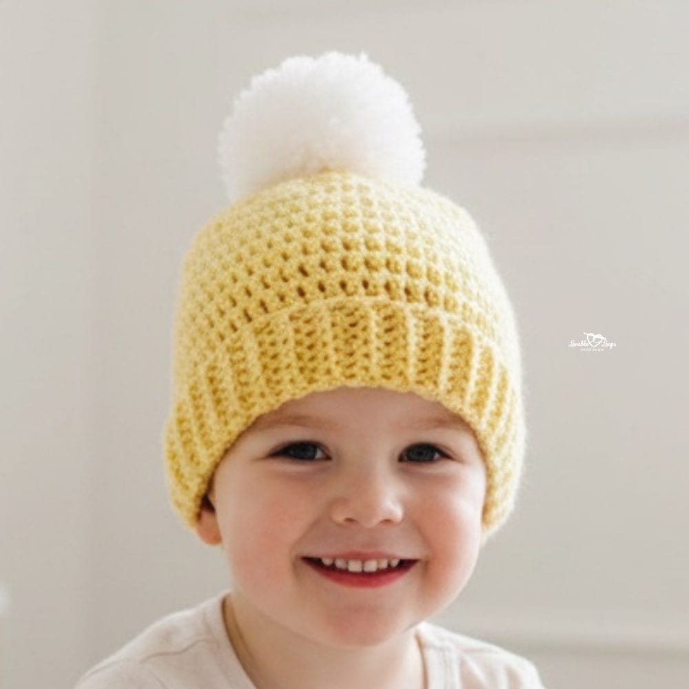 Smiling child wearing a pale yellow crochet hat with a folded brim and large white pom-pom against a soft neutral background.