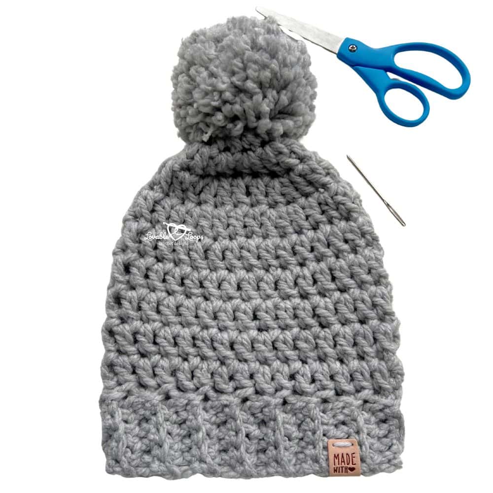 Gray chunky crochet beanie with a ribbed brim and pom pom shown flat with scissors and a yarn needle on a light background.