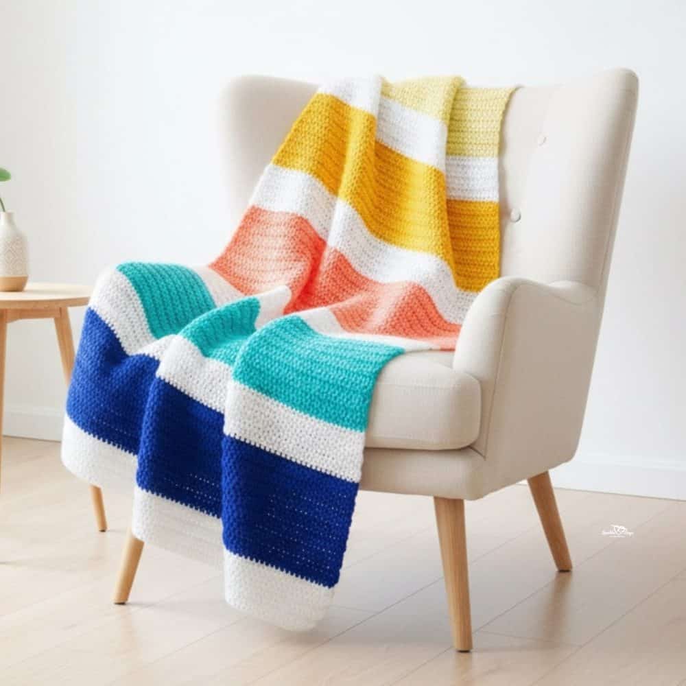 Color block crochet blanket draped over a cream wingback chair in a bright room, featuring wide stripes in yellow, peach, aqua, navy, and white.