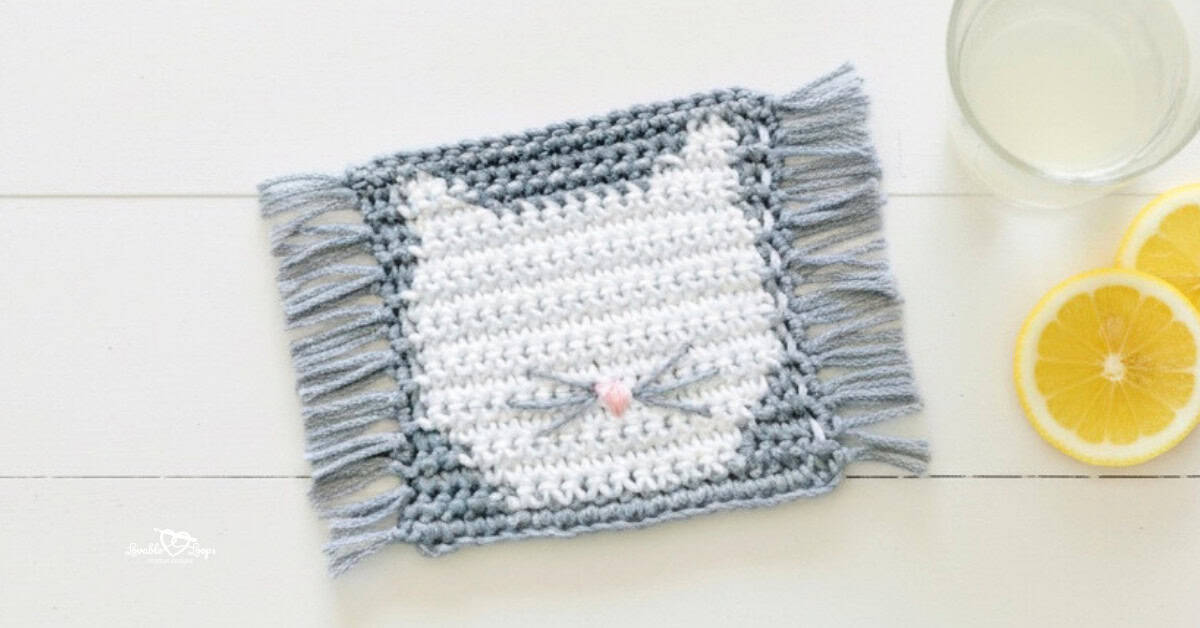 Top-down photo of a gray crochet cat coaster with fringe on both sides, a white cat face, pink nose, and stitched whiskers on a whitewashed table beside a glass of lemonade and lemon slices.
