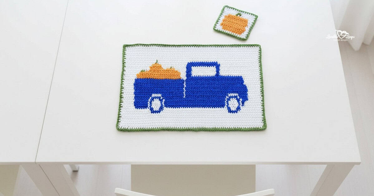 Overhead view of a white crochet placemat on a white table, featuring a blue vintage pickup truck carrying orange pumpkins, with a matching square crochet coaster above it.