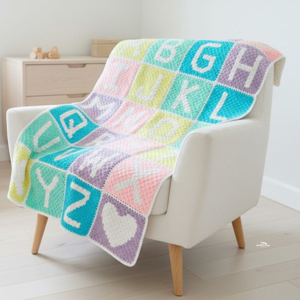 Pastel C2C alphabet crochet blanket draped over a cream armchair, featuring letter blocks in soft baby colors.