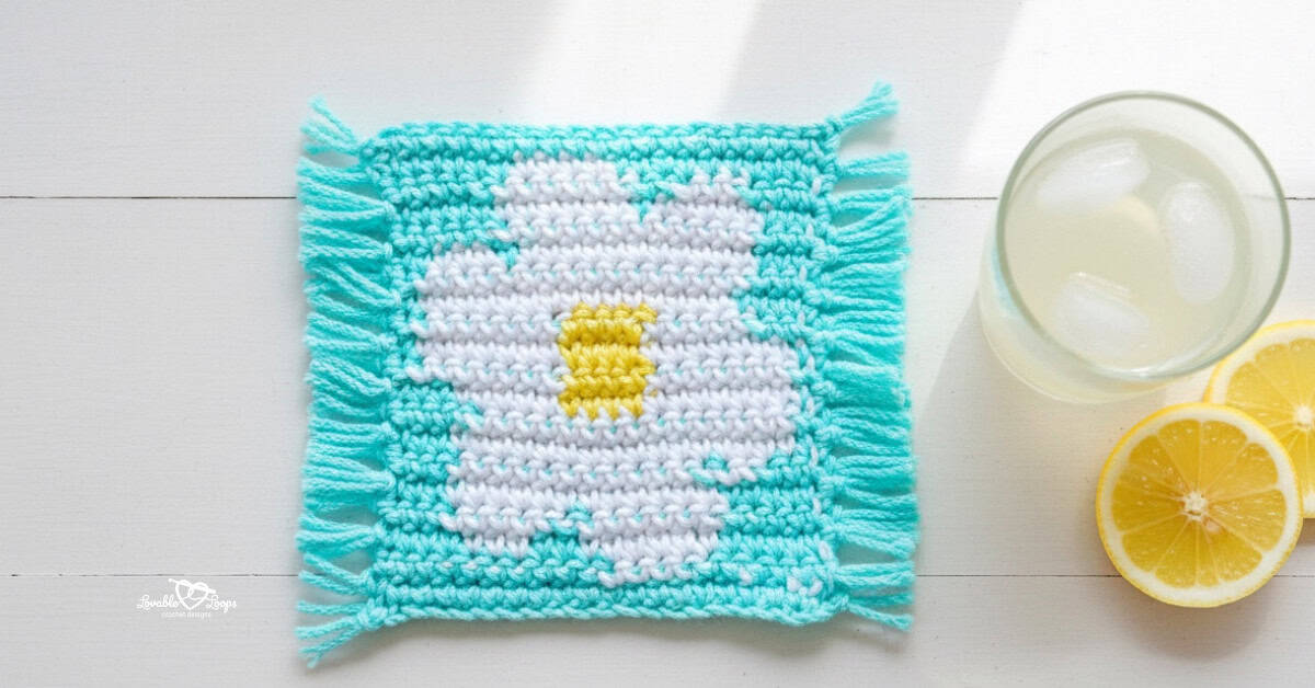 Top-down photo of a turquoise crochet daisy coaster with fringe on both sides, featuring a white flower and yellow center, styled on a whitewashed table beside a glass of lemonade and lemon slices.