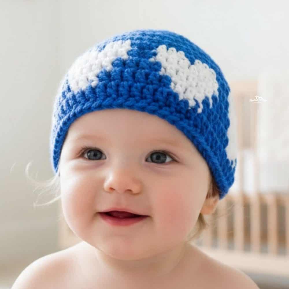 Baby wearing a blue crochet hat with white heart motifs in a bright nursery.