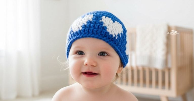 Baby wearing a blue crochet hat with white heart motifs in a soft neutral nursery.
