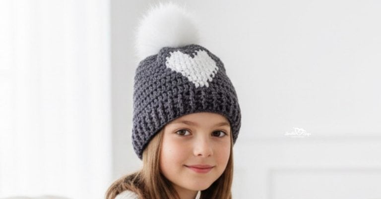 Child wearing a gray crochet heart beanie with a white pom pom in a bright cozy living room with soft neutral decor