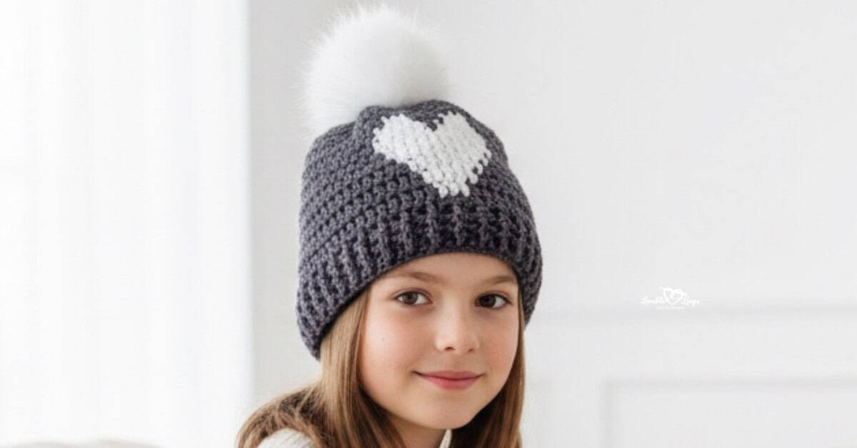 Child wearing a gray crochet heart beanie with a white pom pom in a bright cozy living room with soft neutral decor
