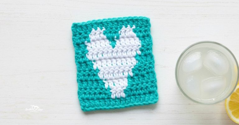 Top-down photo of a teal crochet heart coaster with a white heart design on a whitewashed table beside a glass of lemonade and a lemon slice.