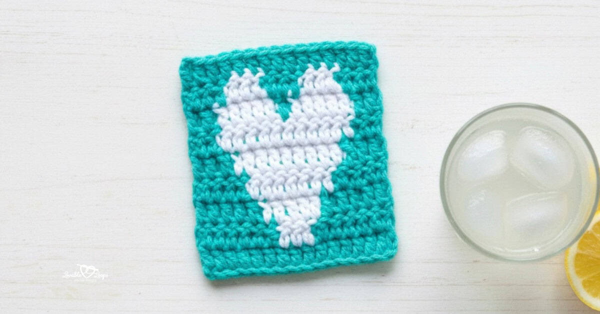 Top-down photo of a teal crochet heart coaster with a white heart design on a whitewashed table beside a glass of lemonade and a lemon slice.