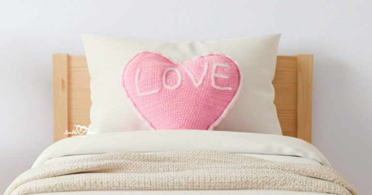 Pink crochet conversation heart pillow with “LOVE” propped on a pine toddler bed with white bedding.