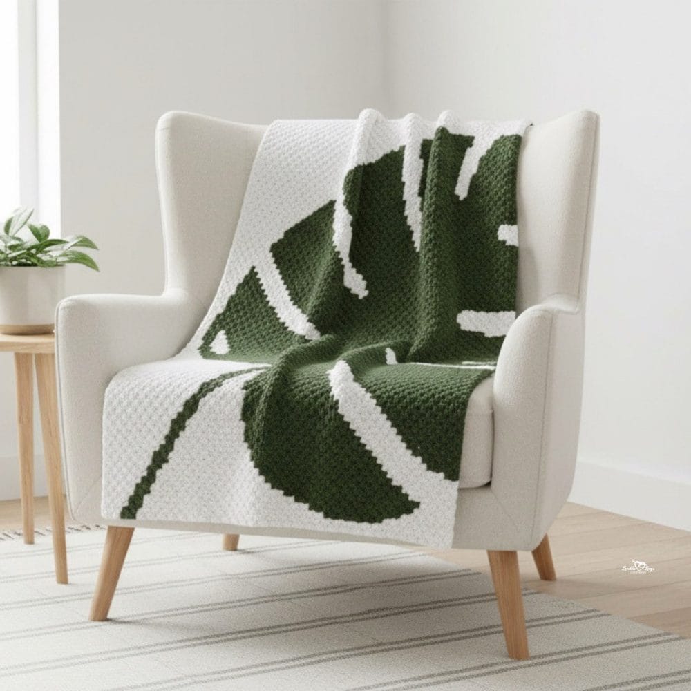 White C2C monstera leaf blanket draped over a cream wingback chair in a bright neutral room, featuring a large dark green leaf design.
