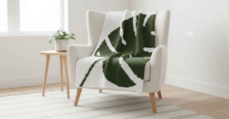 White C2C crochet blanket with a dark green monstera leaf design draped over a cream armchair in a bright room.