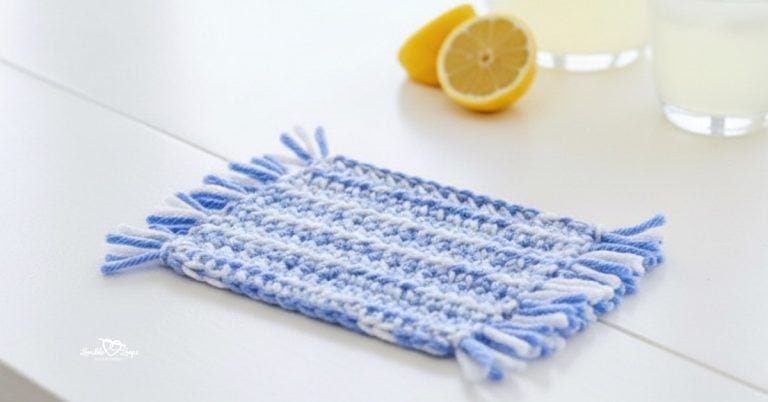 Angled close-up photo of a blue and white striped crochet mug rug with fringe on both short ends, styled on a whitewashed table with lemonade and lemon slices in the background.