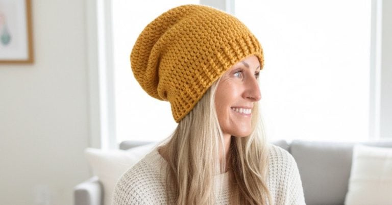 Woman wearing a mustard yellow crochet slouchy beanie and cream sweater, seated in a bright cozy living room with neutral decor and soft natural light.